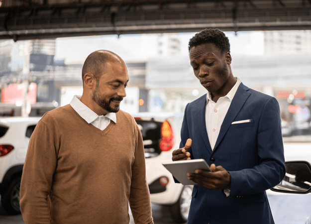 A black man gestures toward a tablet device he's holding as another man standing next to him looks on. They are standing in a dealership showroom.